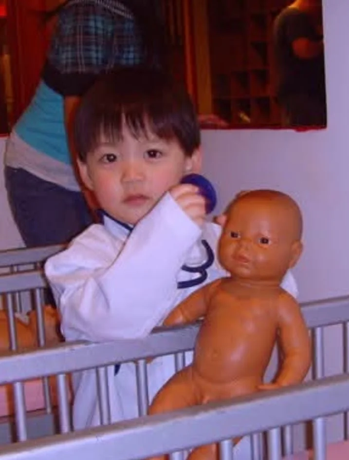 Aaron as a toddler in a white coat with a stethoscope, holding a baby doll