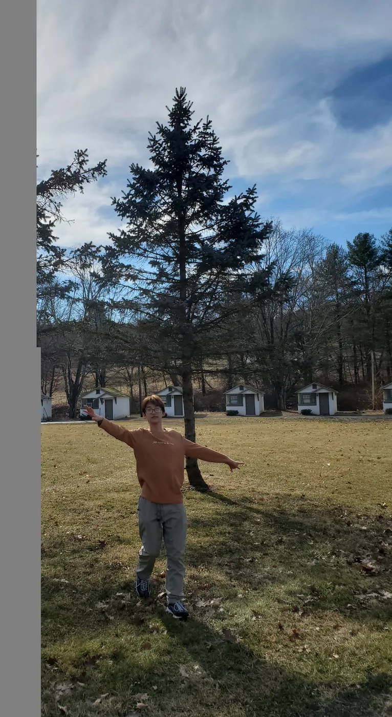 Aaron with arms out in a grassy field by a pine tree